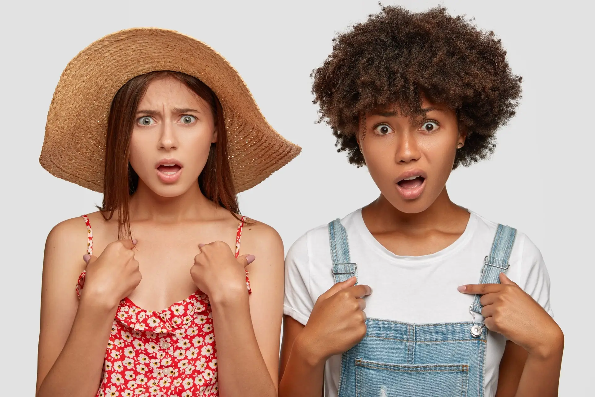 Photo of indignant discontent multiethnic women point at themselves, frown faces in displeasure, have displeased facial expressions, model against white background. Why should we do exactly this?
